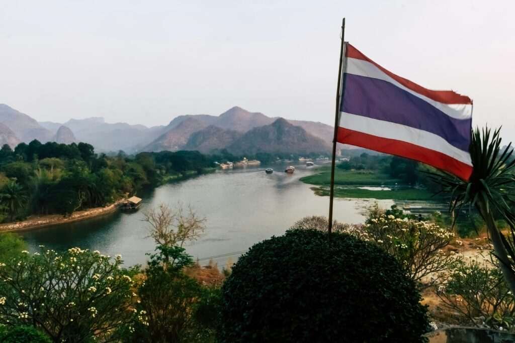 A picturesque river flowing through the lush mountains of Kanchanaburi, Thailand, with a Thai flag in the foreground.